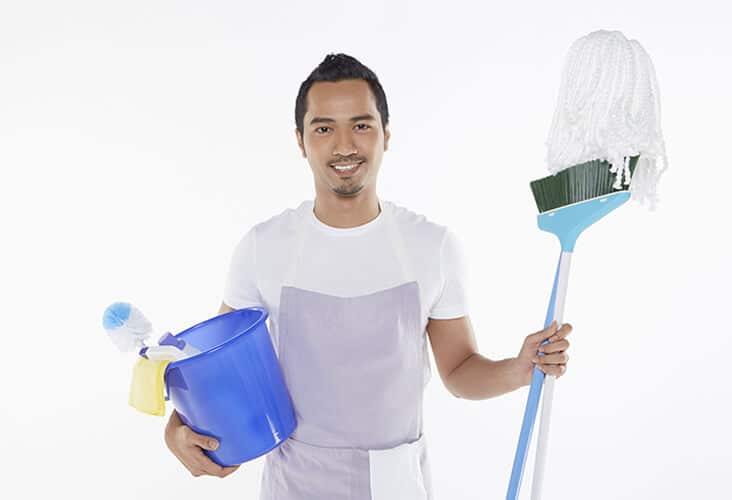 A cleaner stands smiling holding a bucket and brush